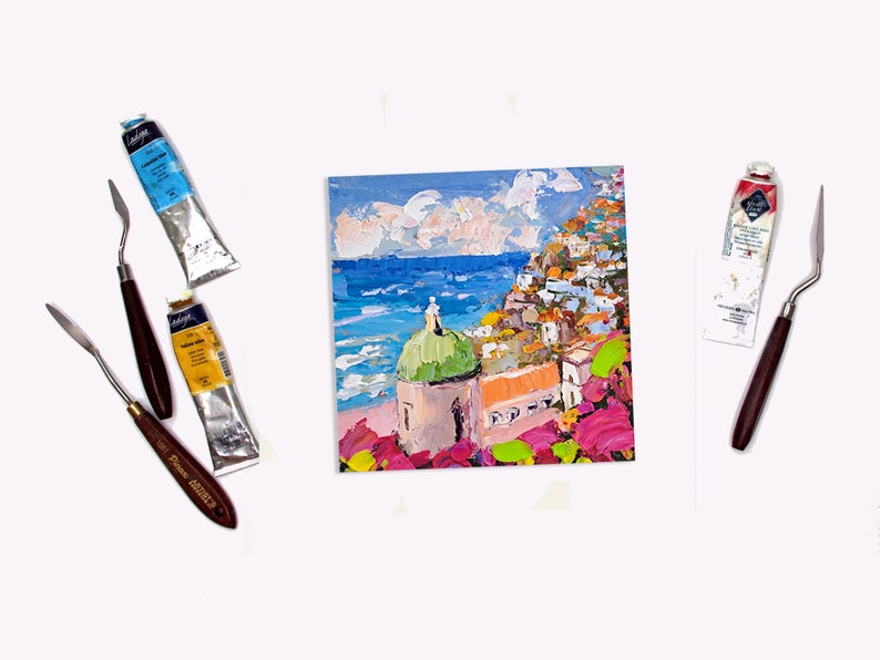 May include: A painting of a colorful Italian village with a green domed church, overlooking a blue ocean. The painting is done in a textured style with visible brushstrokes. The painting is surrounded by paint tubes and palette knives.