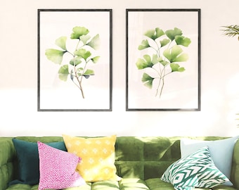 Puede incluir: Dos cuadros de pared con ilustraciones en acuarela de hojas de ginkgo verdes sobre fondo blanco, presentados en marcos negros delgados sobre un sofá de terciopelo verde capitoné con cojines coloridos en amarillo, rosa y azul verdoso.