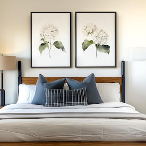 May include: A wooden bed frame with a light-colored comforter and pillows. Two framed floral prints hang above the bed. A lamp with a beige shade sits on a nightstand. The room has a neutral color palette with blue accents.