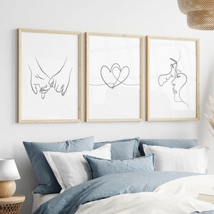 Op de afbeelding: Drie ingelijste kunstdrukken met lijntekeningen. De prints tonen handen die pinken vasthouden, een hart en twee gezichten in profiel. De frames zijn van licht hout en de kunst wordt tentoongesteld boven een bed met blauw en wit beddengoed.
