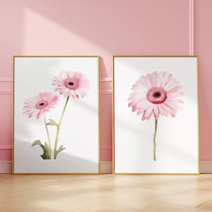May include: Two framed watercolour paintings of pink gerbera daisies on a white background. The paintings are in gold frames and are hanging on a pink wall.