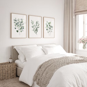 May include: Three framed watercolor prints of green eucalyptus leaves on a white wall above a white bed with a beige knitted throw blanket.