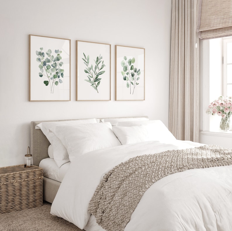 May include: Three framed watercolor prints of green eucalyptus leaves on a white wall above a white bed with a beige knitted throw blanket.