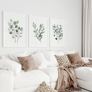 May include: Three framed watercolor prints of green eucalyptus leaves on a white wall above a white sectional sofa with brown throw pillows and a brown throw blanket.