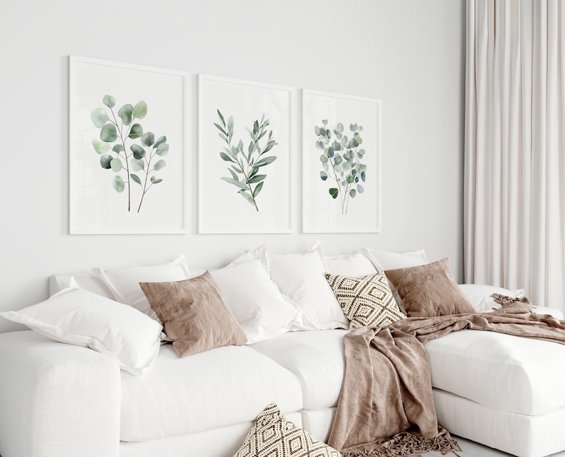 May include: Three framed watercolor prints of green eucalyptus leaves on a white wall above a white sectional sofa with brown throw pillows and a brown throw blanket.