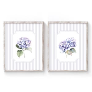 Op de afbeelding: Twee ingelijste prints met aquarel schilderijen van blauwe en paarse hortensia's. De bloemenkunst is geplaatst tegen een wit en grijs gestreepte achtergrond, met een licht houten frame. De botanische kunst is perfect voor woondecoratie.