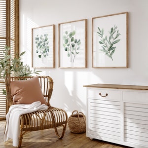 May include: Three framed watercolor prints of green foliage on a white wall. The prints are in wooden frames and are arranged in a row. A wicker chair with a brown cushion and a white throw blanket is in the foreground. A white cabinet with a wooden top is in the background.