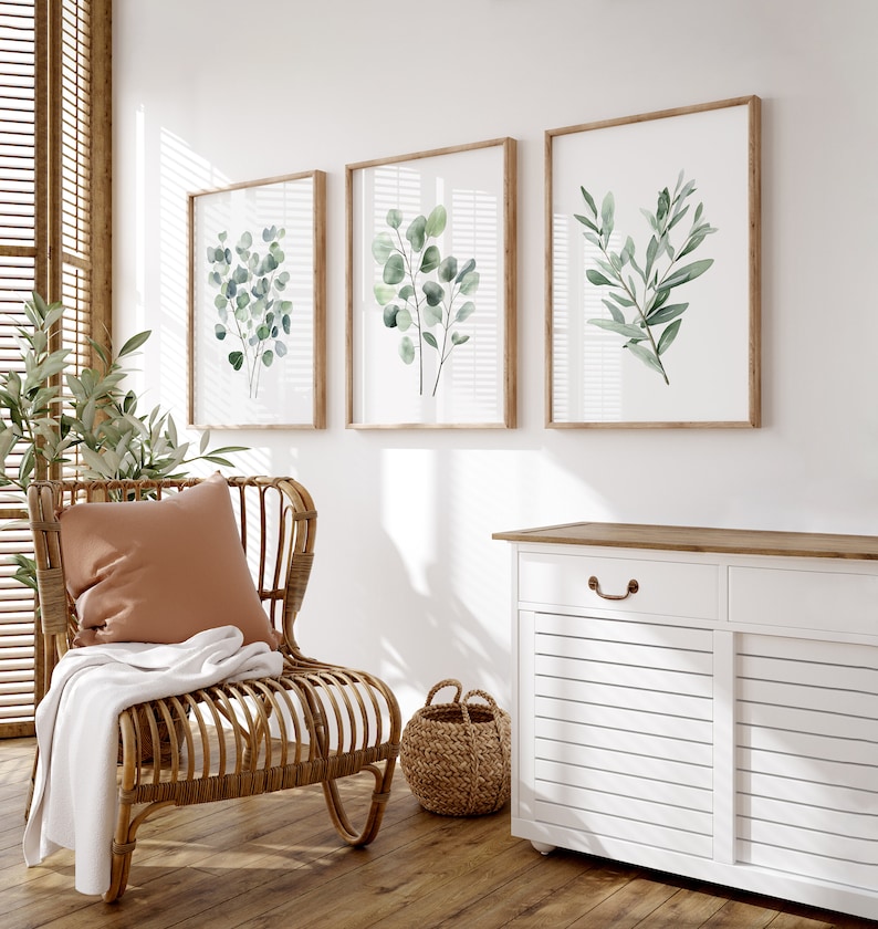 May include: Three framed watercolor prints of green foliage on a white wall. The prints are in wooden frames and are arranged in a row. A wicker chair with a brown cushion and a white throw blanket is in the foreground. A white cabinet with a wooden top is in the background.
