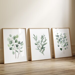 May include: Three framed watercolor prints of green eucalyptus leaves on a white background. The frames are light wood and the prints are arranged in a row.