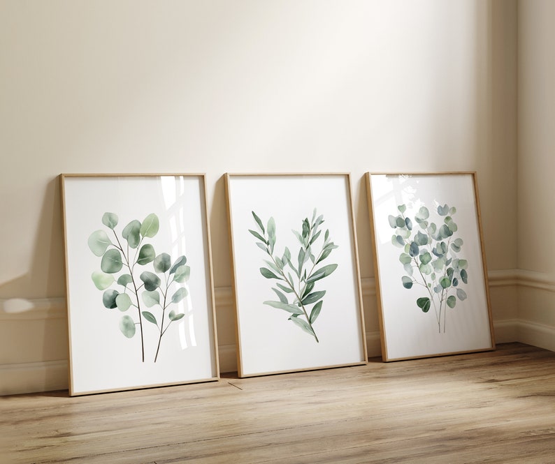 May include: Three framed watercolor prints of green eucalyptus leaves on a white background. The frames are light wood and the prints are arranged in a row.
