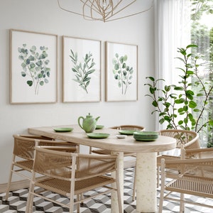 May include: A dining room with a wooden table and six chairs. The table is oval shaped and has a light wood finish. The chairs are made of woven wicker and have a natural brown color. There are three framed prints on the wall behind the table, each featuring a watercolor painting of green leaves. The room has a white tiled floor with a black and white geometric pattern.