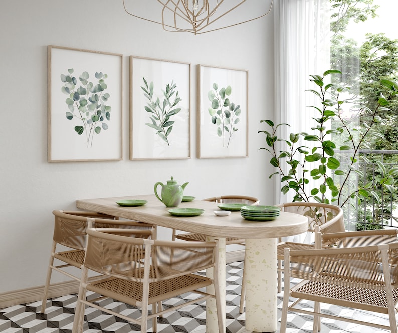 May include: A dining room with a wooden table and six chairs. The table is oval shaped and has a light wood finish. The chairs are made of woven wicker and have a natural brown color. There are three framed prints on the wall behind the table, each featuring a watercolor painting of green leaves. The room has a white tiled floor with a black and white geometric pattern.