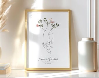 May include: A gold framed print featuring a black line drawing of two hands clasped together. The hands are surrounded by small green leaves and pink flowers. The text "Anna & Nicolas You Are My Person 02.21.2023" is printed below the drawing.