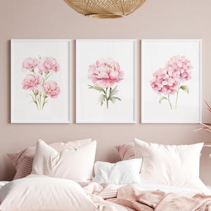 Pode incluir: Três impressões de arte floral emolduradas com pinturas em aquarela de flores rosa. As impressões são exibidas acima de uma cama com roupa de cama e travesseiros rosa. As flores incluem cravos, uma peônia e hortênsias.