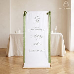 May include: A white wedding welcome sign with green ribbon accents. The sign features the names "Ashley & Meine" and the date "October 25, 2026". The sign is framed in gold and stands on a wooden floor.