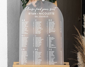 May include: A white acrylic arch-shaped sign with a seating chart for a wedding. The sign has the text "Please find your seat" at the top, followed by "Ryan + Nicolette" and the date "06.25.2023". The sign lists guest names in alphabetical order.