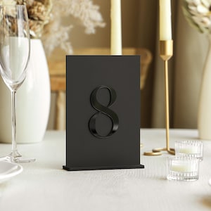 May include: A black table number eight, displayed on a white tablecloth. The number is in a bold, elegant font. The scene includes champagne flutes, candles, and floral arrangements, suggesting a wedding or event setting.