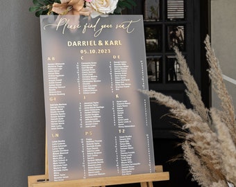 Puede incluir: Un cartel de asignación de asientos de acrílico gris con letras doradas para una recepción de boda. El cartel enumera los nombres de los invitados y sus mesas asignadas. El cartel está en un caballete de madera y decorado con flores y hierbas secas.