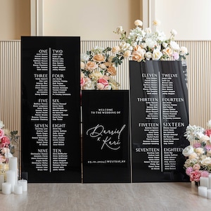 May include: Black acrylic wedding seating chart signs with white text. The signs are arranged in two columns, with the names of guests listed under each table number. The signs are decorated with floral arrangements.