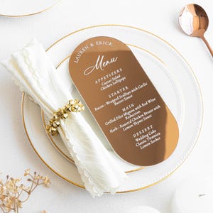 May include: A gold-colored menu card with rounded edges, featuring the names "Lauren & Erick" and a list of menu items. The card rests on a white plate with a gold rim, next to a folded white napkin and a gold napkin ring.