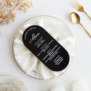 May include: A black, arched menu card with white text, featuring a wedding menu. The menu is on a white plate with a gold rim, accompanied by gold cutlery. The menu lists appetizers, starters, main courses, and desserts.