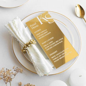 Könnte beinhalten: Eine goldfarbene Menükarte mit dem Text "Food Menu" und einer Liste von Gerichten, platziert auf einem weißen Teller mit Goldrand. Eine weiße Serviette mit einem goldenen Ring und ein goldener Löffel sind ebenfalls zu sehen.