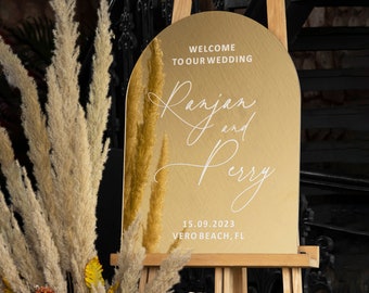 May include: Gold arch-shaped wedding sign with the text "Welcome to our wedding Ranjan and Perry 15.09.2023 Vero Beach, FL" in white lettering.
