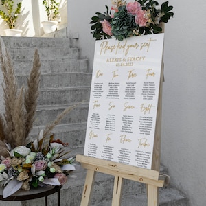 K&ouml;nnte beinhalten: Eine wei&szlig;e Hochzeits-Sitzordnung aus Acryl mit goldfarbenen Buchstaben, die auf einer h&ouml;lzernen Staffelei pr&auml;sentiert wird. Die Tabelle listet Namen unter den Nummern eins bis zehn auf, mit den Namen "ALEXIS & STACEY" und dem Datum "03.04.2023" oben. Blumengestecke schm&uuml;cken die Oberseite und den Sockel.