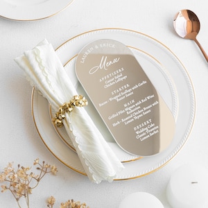 May include: A wedding menu card with a gold-toned finish, featuring a list of appetizers, main courses, and desserts. The menu is placed on a white plate with a gold rim, next to a folded white napkin and a gold napkin ring.