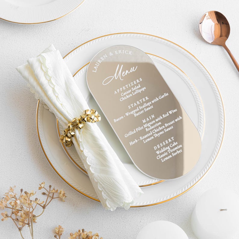May include: A wedding menu card with a gold-toned finish, featuring a list of appetizers, main courses, and desserts. The menu is placed on a white plate with a gold rim, next to a folded white napkin and a gold napkin ring.
