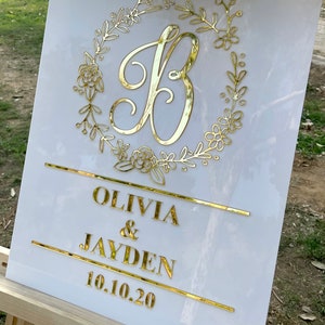 May include: White acrylic wedding sign with gold lettering. The sign features a floral wreath with the letter "B" in the center. Below the wreath, the names "Olivia & Jayden" and the date "10.10.20" are written.