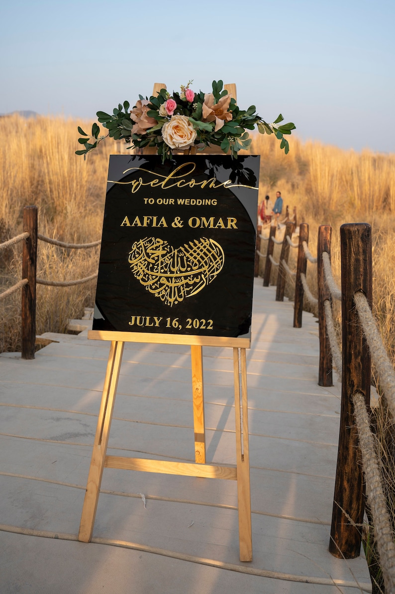 May include: A black welcome sign with gold lettering announcing the wedding of Aafia & Omar on July 16, 2022. The sign is decorated with a gold heart featuring Arabic calligraphy and is displayed on a wooden easel.