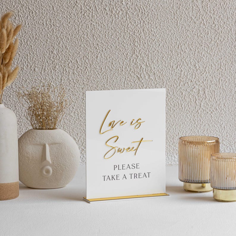 Peut inclure: Panneau en acrylique blanc avec l'inscription dor&eacute;e &laquo; Love is Sweet, Please Take a Treat &raquo;. Le panneau est pos&eacute; sur un support dor&eacute;. Deux bougeoirs en verre ambr&eacute; sont &agrave; droite. Fond textur&eacute;.
