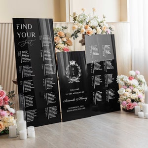 May include: Black acrylic wedding seating chart signs with white text. The signs are arranged in a row on a wooden floor. The signs include the names of guests and table numbers. The signs also include the text "Find Your Seat", "Welcome to the Wedding of Amanda & Henry", "October 15, 2024", and "Highland, Indiana".