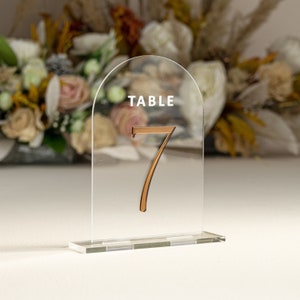 May include: Clear acrylic table number holder with a rounded arch top. The number "7" is printed in a copper color on the front of the holder. The text "TABLE" is printed in white at the top of the holder.