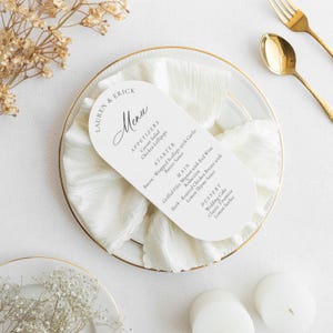 May include: A white menu card with rounded edges, featuring the names "LAUREN & ERICK" and the word "Menu" in elegant script. The menu lists appetizers, a starter, main courses, and dessert options. The card rests on a white napkin and a gold-rimmed plate.