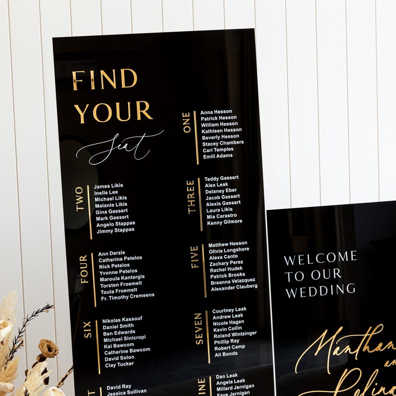 May include: Black acrylic seating chart with gold lettering. The chart is divided into nine sections, each with a number and a list of names. The text "Find Your Seat" is at the top of the chart. The text "Welcome to Our Wedding" is at the bottom of the chart.