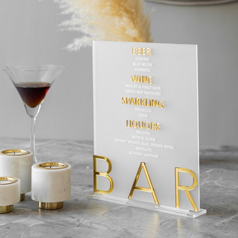 May include: A white acrylic sign with gold lettering that reads "BEER", "WINE", "SPARKLING", "LIQUORS", and "BAR". The sign lists different types of beer, wine, sparkling wine, and liquor.