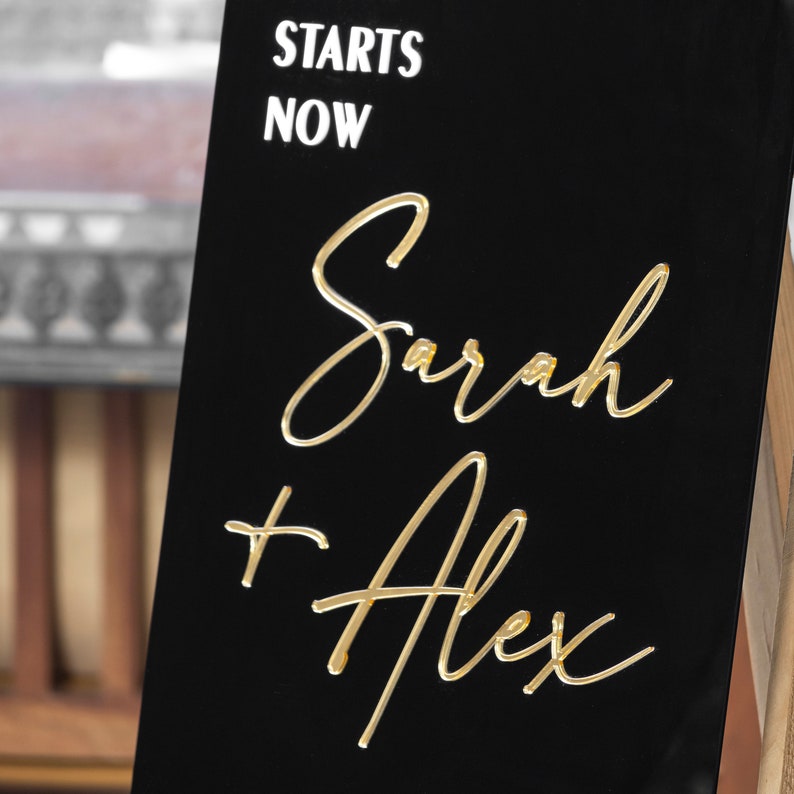 May include: Black sign with white text "Starts Now" and gold lettering "Sarah + Alex" on a wooden easel.