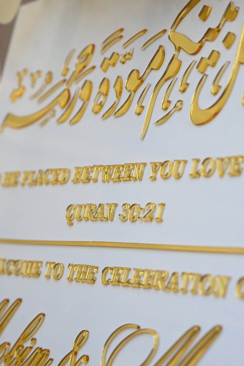 Puede incluir: Un cartel blanco con letras doradas que dice "To be placed between you love Quran 30:21" y "Welcome to the celebration of" con una escritura cursiva dorada en la parte inferior.