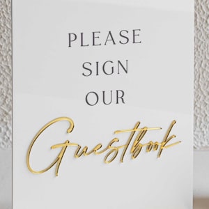 Peut inclure: Panneau blanc avec l'inscription "PLEASE SIGN OUR Guestbook" en lettres noires et dor&eacute;es. L'&eacute;criture dor&eacute;e est en cursive. Le panneau repose sur un socle dor&eacute;.