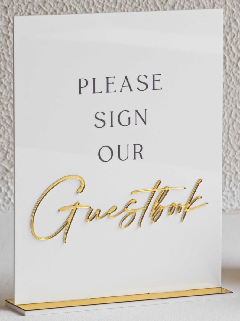 Peut inclure: Panneau blanc avec l'inscription "PLEASE SIGN OUR Guestbook" en lettres noires et dor&eacute;es. L'&eacute;criture dor&eacute;e est en cursive. Le panneau repose sur un socle dor&eacute;.