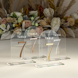 May include: Four clear acrylic table number holders with a rounded arch top. Each holder has a white "TABLE" inscription and a gold or rose gold number "7".