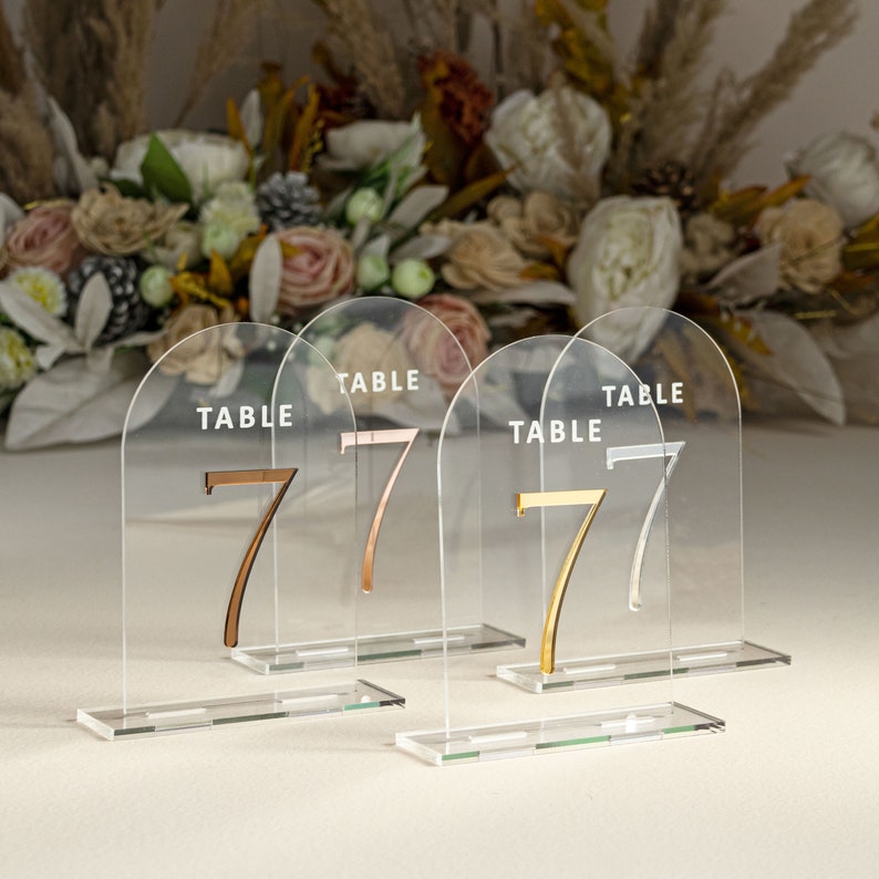 May include: Four clear acrylic table number holders with a rounded arch top. Each holder has a white "TABLE" inscription and a gold or rose gold number "7".