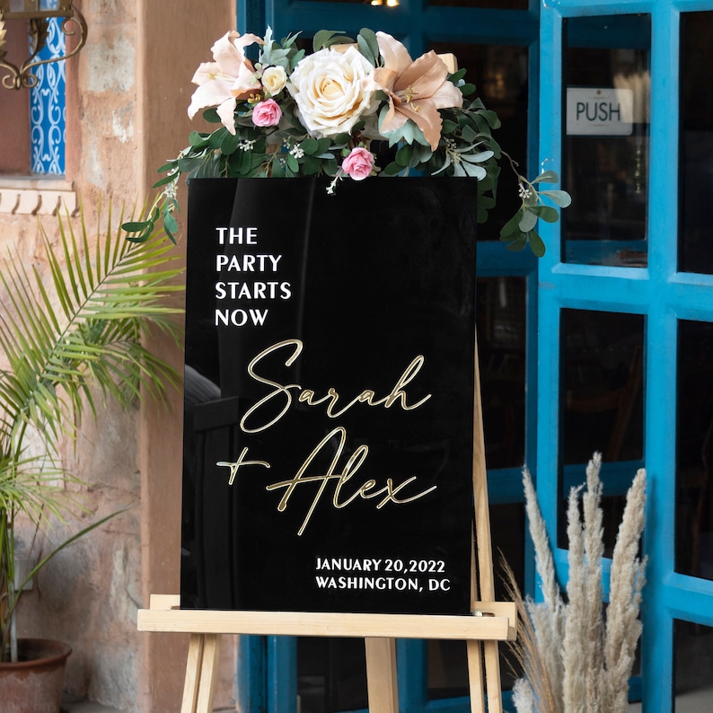 May include: Black acrylic wedding sign with gold lettering. The sign reads "The Party Starts Now" followed by "Sarah + Alex" and the date "January 20, 2022" and location "Washington, DC".