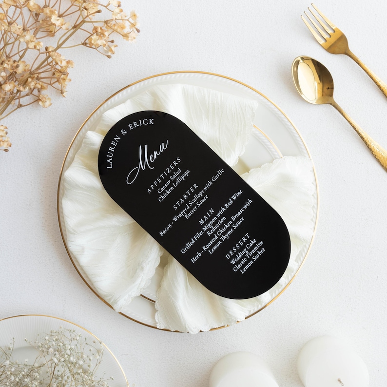 May include: A black, arched menu card with white text, featuring a wedding menu. The menu is on a white plate with a gold rim, accompanied by gold cutlery. The menu lists appetizers, starters, main courses, and desserts.