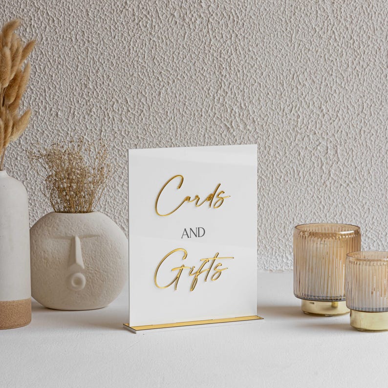 Peut inclure: Panneau en acrylique blanc avec l'inscription dor&eacute;e "Cards and Gifts". Le panneau est pos&eacute; sur un socle dor&eacute;. Deux bougeoirs en verre ambr&eacute; avec des bases dor&eacute;es sont &agrave; droite.