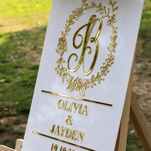 May include: White wedding sign with gold lettering. The sign features a gold letter "B" surrounded by a floral wreath. The text below reads "Olivia & Jayden 10.10.20".