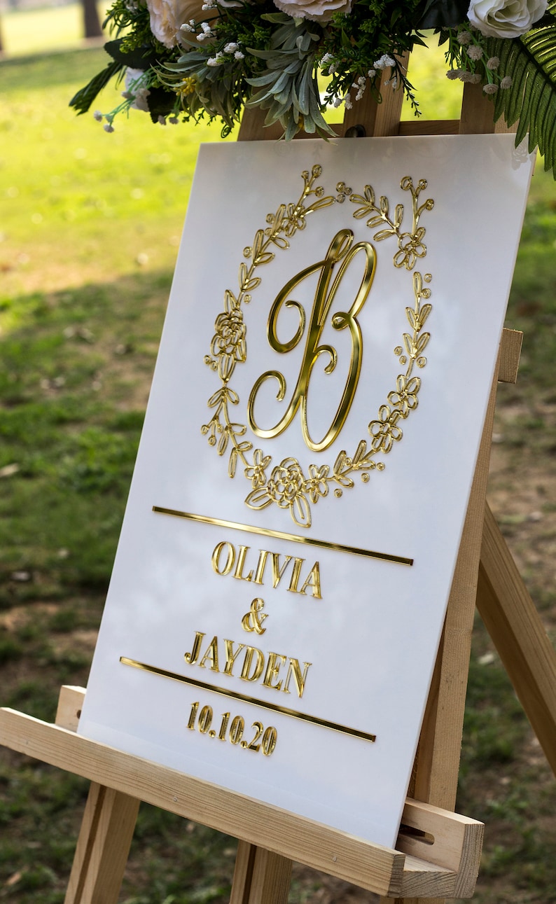 May include: White wedding sign with gold lettering. The sign features a gold letter "B" surrounded by a floral wreath. The text below reads "Olivia & Jayden 10.10.20".