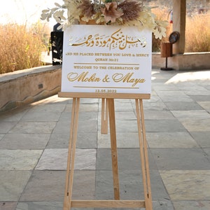Puede incluir: Un cartel blanco con letras doradas en un caballete. El cartel dice "And He Placed Between You Love & Mercy, Quran 30:21, Welcome to the Celebration of Mobin & Maya, 12.05.2022".